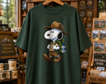 Könnte beinhalten: Dunkelgrünes T-Shirt mit Snoopy als Park Ranger, der einen braunen Hut, ein khakifarbenes Hemd trägt und einen Rucksack und eine Wasserflasche trägt. Das T-Shirt hängt an einem Holzbügel. Die Worte "EXPLORE DREAM DISCOVER" sind im Hintergrund zu sehen.