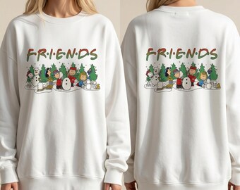 Könnte beinhalten: Ein weißes langärmeliges Sweatshirt mit Rundhalsausschnitt, das den Schriftzug FRIENDS in roten und grünen Buchstaben über einer grafischen Illustration von Peanuts-Figuren zeigt, die im Schnee mit Tannenbäumen und Schneemännern spielen.