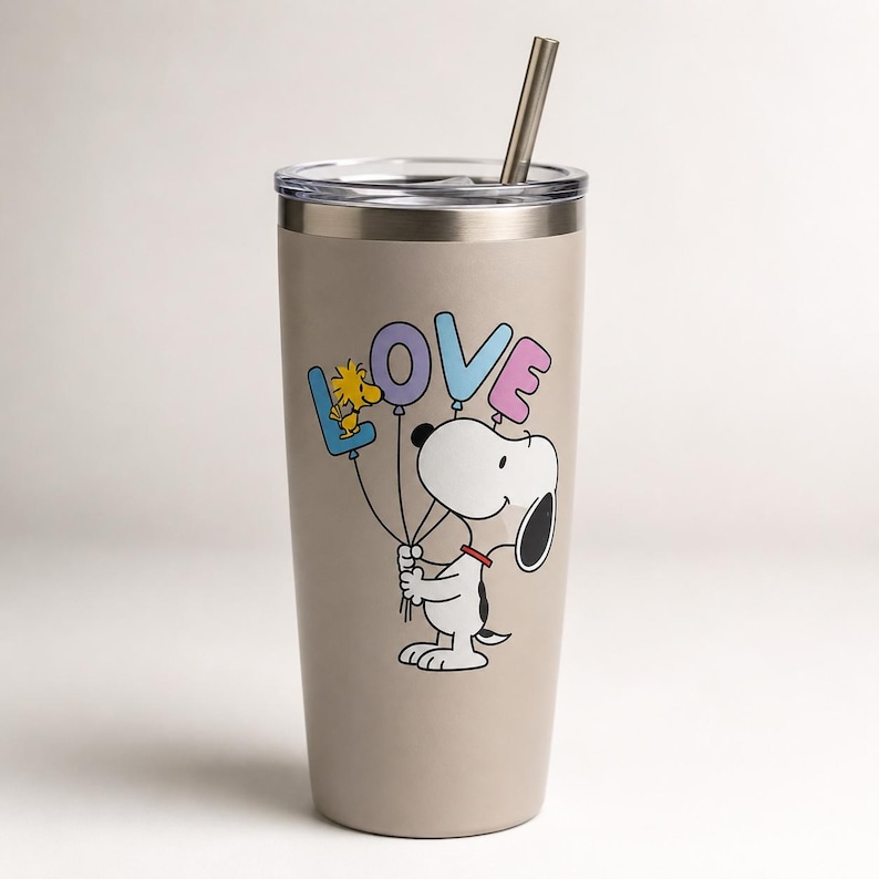 May include: A beige stainless steel tumbler with a clear lid and a metal straw. The tumbler features a graphic of Snoopy holding four balloons shaped like the letters L, O, V, and E, with Woodstock perched on the blue L balloon.