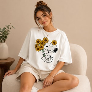 May include: White t-shirt featuring a cartoon Snoopy character holding sunflowers. The graphic is in black and yellow. The shirt has a crew neck and short sleeves. The overall style is casual and relaxed.
