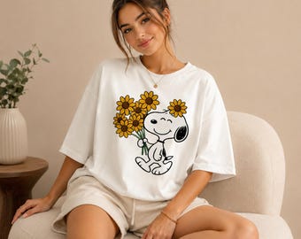 Könnte beinhalten: Weißes T-Shirt mit einem Cartoon-Snoopy-Design, das Sonnenblumen hält. Die Grafik ist schwarz und gelb. Das T-Shirt hat einen Rundhalsausschnitt und kurze Ärmel. Der Stil ist lässig und entspannt.