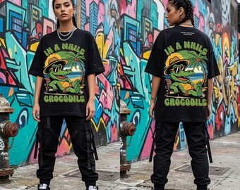 May include: A black short-sleeve t-shirt featuring a graphic of a green crocodile wearing a hat and sunglasses while relaxing on a beach chair under a palm tree. The text "IN A WHILE CROCODILE" is printed in green around the illustration.