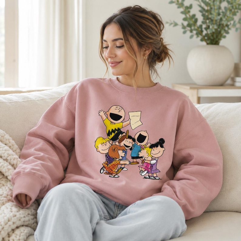 May include: A dusty rose crewneck sweatshirt featuring a colourful graphic print of the Peanuts gang characters, including Charlie Brown, Lucy, Linus, and others, gathered together in a cheerful scene on the front centre of the garment.