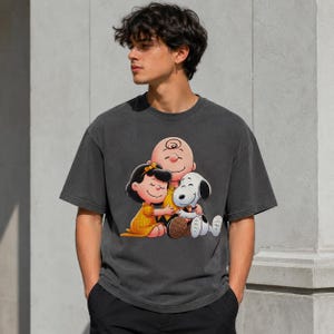 May include: A charcoal grey short-sleeve t-shirt featuring a graphic print of Charlie Brown, Lucy, and Snoopy hugging. The characters are rendered in their classic cartoon style with Lucy wearing a yellow dress and Charlie Brown in a yellow shirt.