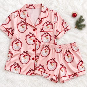 Può includere: Un set di pigiama rosa chiaro composto da una camicia a maniche corte abbottonata con colletto e taschino sul petto, abbinata a pantaloncini coordinati. Il tessuto presenta un motivo ripetuto di volti di Babbo Natale incorniciati da fiocchi rossi.