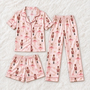 May include: A three-piece pajama set in light pink featuring a repeating pattern of ballerinas in pink tutus, nutcracker soldiers, small bows, and gold stars. The set includes a short-sleeved button-down shirt with a chest pocket, shorts, and pants.