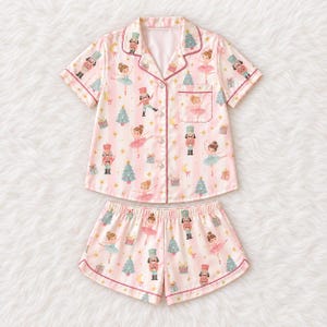 May include: A short-sleeved button-down pyjama shirt and matching shorts set in light pink with vertical white stripes. The fabric features a repeating pattern of nutcrackers, ballerinas, Christmas trees, drums, gift boxes, and yellow crescent moons.