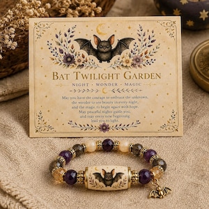 May include: A beaded bracelet featuring purple and translucent beads, gold-toned spacers, and a central cylindrical bead with a bat illustration. A small gold-toned bat charm hangs from the bracelet, which is displayed with a card titled Bat Twilight Garden.
