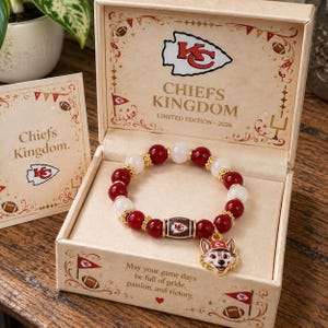 May include: A beaded bracelet featuring alternating red and white beads, gold-tone spacers, a football-shaped bead with the KC logo, and a wolf mascot charm wearing a red cap. The bracelet is presented in a cream-colored box labeled Chiefs Kingdom.