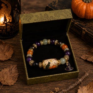 Peut inclure: Un bracelet de perles composé de pierres précieuses violettes, orange et vert pâle séparées par des entretoises en métal doré. Une perle cylindrique centrale représente un chat noir, des citrouilles et un croissant de lune. Une breloque chapeau de sorcière dorée y est suspendue.