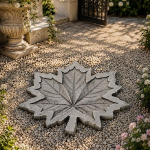 May include: A grey concrete maple leaf stepping stone, with detailed veining and a textured surface. The stone is set on a bed of small pebbles. The background features a garden and gate.