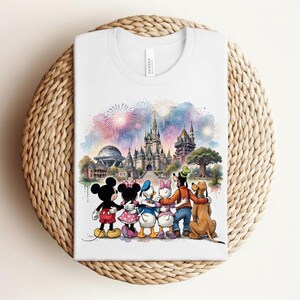 May include: White t-shirt featuring a watercolor illustration of Disney characters in front of a castle and fireworks display. Mickey Mouse, Minnie Mouse, Donald Duck, Daisy Duck, Goofy, and Pluto are depicted. The shirt is folded on a woven surface.