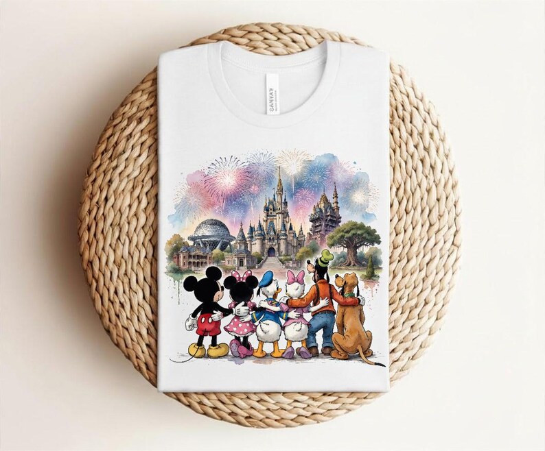 May include: White t-shirt featuring a watercolor illustration of Disney characters in front of a castle and fireworks display. Mickey Mouse, Minnie Mouse, Donald Duck, Daisy Duck, Goofy, and Pluto are depicted. The shirt is folded on a woven surface.