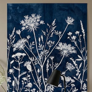 Pode incluir: Uma grande impressão de arte de parede retangular com um design botânico de flores silvestres brancas sobre um fundo azul escuro. A obra de arte é exibida acima de uma mesa de cabeceira de madeira com uma lâmpada e um livro.