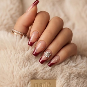 Könnte beinhalten: Lange, sargförmige Fingernägel mit tiefroten French-Tips auf nudefarbener Basis. Die Nägel sind mit kleinen, klaren Strasssteinen und einem blumenförmigen Kristall verziert. Eine goldene Plakette zeigt den Schriftzug nailè Atelier.