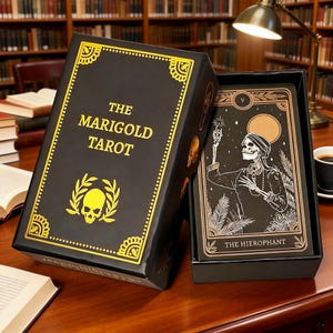 May include: A black tarot card box set with gold detailing, labelled "THE MARIGOLD TAROT". The box has a skull and laurel wreath emblem. A card is visible, featuring a skeleton illustration and the words "THE HIEROPHANT".