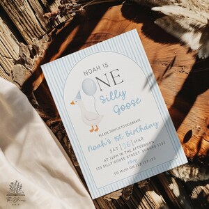 May include: A rectangular birthday invitation card with a light blue vertical striped border. The text reads: Noah is ONE silly goose. Please join us to celebrate Noah's 1st birthday. Sat 26 Mar at 12pm in the afternoon, 123 Silly Goose Street, Suburb 1234. RSVP to Mum on 123 123 123. A white goose wearing a party hat and holding a blue balloon is illustrated inside an arched frame.