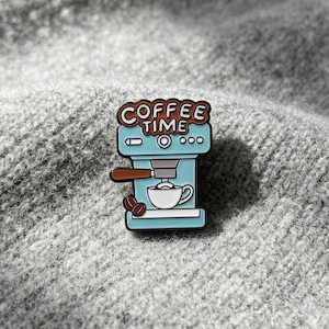 Puede incluir: Un pin de esmalte azul claro con forma de máquina de espresso con un mango marrón y dos granos de café. El texto COFFEE TIME está escrito en letras marrones en la parte superior. Una taza de café blanca se sitúa bajo el portafiltro.
