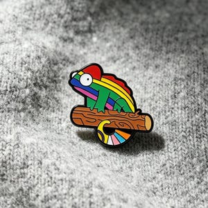 May include: A metal enamel pin shaped like a chameleon with a rainbow-striped body, a large white eye, and a curled tail, perched on a brown wooden branch with black outlines and a smooth, glossy finish, featuring vibrant red, yellow, and green colors.