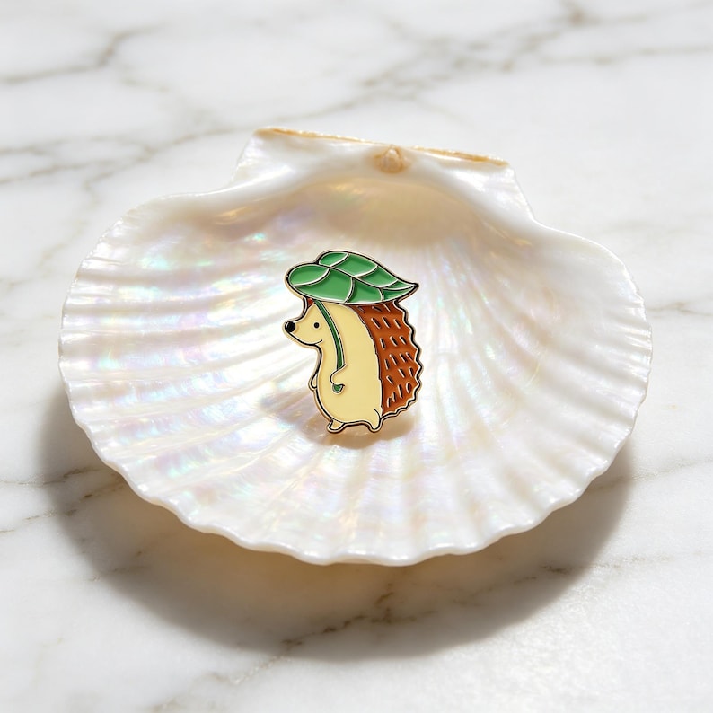 May include: A cute enamel pin of a hedgehog wearing a green leaf hat. The hedgehog has a tan body and a brown spiky back. The pin is set on a white seashell. A delightful accessory.