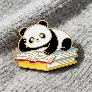 Pode incluir: Um broche de esmalte metálico com um panda preto e branco apoiando a cabeça em uma pata enquanto está deitado sobre uma pilha de três livros. Os livros têm capas azul-claras, brancas e amarelas com páginas visíveis e contorno dourado.