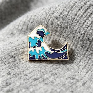 May include: A gold-rimmed enamel pin shaped like a stylised ocean wave. The design features layered sections of deep navy blue, bright turquoise, and white enamel, creating a graphic representation of a cresting wave with a smooth, polished finish.