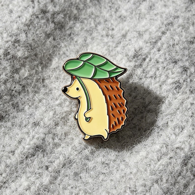 May include: An enamel pin depicting a cartoon hedgehog with a green leaf hat. The hedgehog has a tan body, brown quills, and a black nose. The pin is set against a grey background.