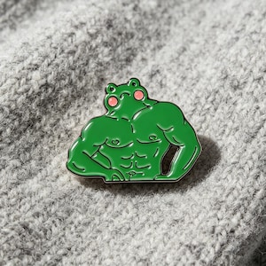 Könnte beinhalten: Ein grüner Emaille-Pin in Form eines muskulösen Frosches mit definierten Brust- und Bauchmuskeln, rosa runden Wangen und einem neutralen Gesichtsausdruck. Der Pin hat schwarze Umrandungen und eine glatte, glänzende Oberfläche.