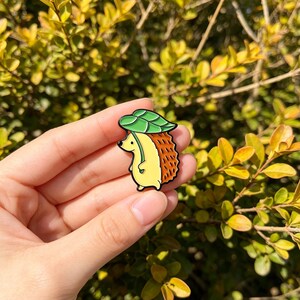 May include: A colourful enamel pin depicting a cartoon hedgehog with a green leaf hat. The hedgehog has a yellow body, brown spiky back, and a green leaf hat. The pin is set against a backdrop of green and yellow foliage.