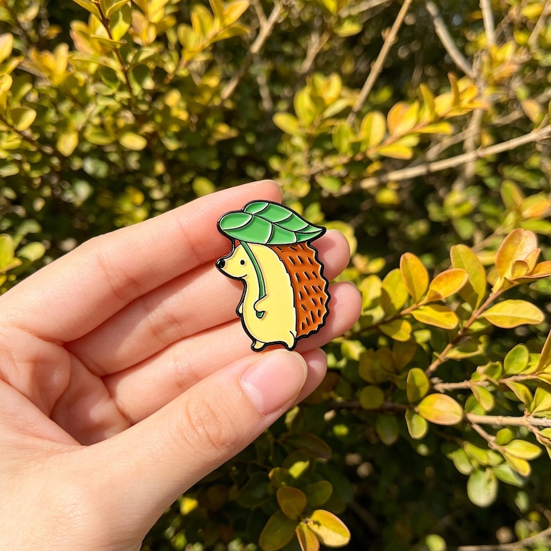 May include: A colourful enamel pin depicting a cartoon hedgehog with a green leaf hat. The hedgehog has a yellow body, brown spiky back, and a green leaf hat. The pin is set against a backdrop of green and yellow foliage.