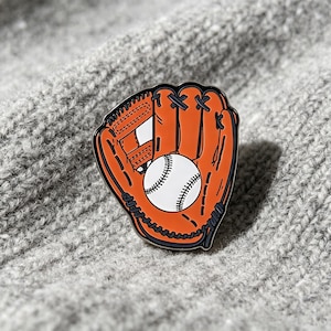Könnte beinhalten: Eine Emaille-Anstecknadel in Form eines Baseballhandschuhs in leuchtendem Orange mit schwarzen Umrandungen. Der Handschuh zeigt einen weißen Baseball mit schwarzen Nähten in der Mitte der Handfläche. Das Design enthält detaillierte schwarze Linien.