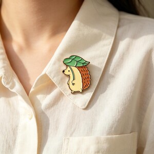 May include: An enamel pin depicting a hedgehog wearing a leaf hat. The pin has a yellow body, brown quills, and a green leaf. It is attached to a cream-coloured shirt.