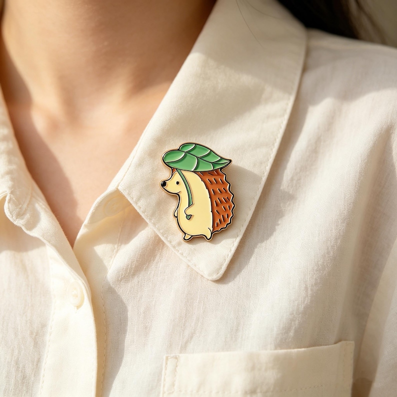May include: An enamel pin depicting a hedgehog wearing a leaf hat. The pin has a yellow body, brown quills, and a green leaf. It is attached to a cream-coloured shirt.