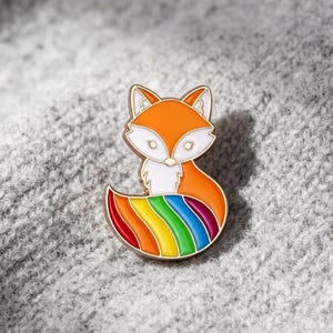 May include: A metal enamel pin shaped like a fox with orange and white fur on its head and chest. The fox has a large, curved tail featuring horizontal stripes in rainbow colors including red, orange, yellow, green, blue, and purple.