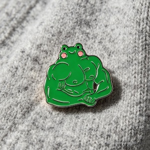 May include: A green enamel pin shaped like a muscular frog with a smiling face and pink cheeks. The frog has defined chest and arm muscles, with black outlines and a metallic border. The design features a bright green body with small pink circles on the cheeks and black dots for eyes, showcasing a humorous and stylized character illustration.