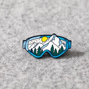 Puede incluir: Un pin de esmalte metálico con forma de gafas de esquí con montura azul. La lente presenta una ilustración gráfica de montañas blancas nevadas, un sol amarillo brillante y pinos verde oscuro sobre un cielo azul claro con puntos negros.