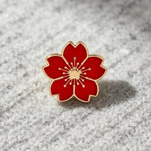 Puede incluir: Un broche de esmalte con forma de flor de cinco pétalos, con un relleno de esmalte rojo vibrante y un contorno de metal dorado. El centro de la flor muestra delicados estambres dorados que irradian hacia afuera, creando un diseño botánico.