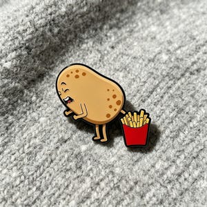 May include: A tan enamel pin featuring a cartoon potato character with brown spots and a facial expression of distress, standing next to a small red container filled with golden yellow french fries, outlined in black metal.