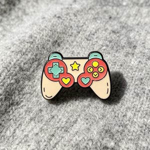 Op de afbeelding: Een metalen emaille speld in de vorm van een videogamecontroller. De controller is crèmekleurig met rode accenten, voorzien van een mintgroene richtingsknop, een gele ster, een geel hartje, een mintgroen hartje en vier gele ronde knoppen.