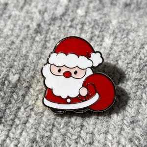Può includere: Una spilla in smalto metallico raffigurante un Babbo Natale in stile cartoon che indossa un cappotto e un cappello rossi con bordi bianchi. Babbo Natale ha una grande barba bianca, un naso rosso rotondo e tiene un grande sacco rosso.