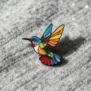 Op de afbeelding: Een kolibrie emaille speld met een glas-in-loodstijl ontwerp met levendige segmenten in blauw, geel, oranje, groen, rood en paars. De vogel is afgebeeld in vlucht met gespreide vleugels, met een scherpe snavel en een veelkleurige staart.