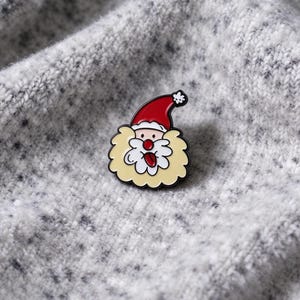 May include: A metal enamel pin featuring a cartoon Santa Claus face with a red hat, a white pom-pom, a large cream-coloured beard, a pink nose, and a small tongue sticking out. The design is outlined in black with a smooth, glossy finish.