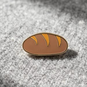 May include: An oval-shaped enamel pin featuring a brown loaf of bread design with three distinct yellow-orange diagonal score marks across the top surface, all outlined in a thin gold-toned metal border.