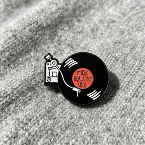 May include: A black and white enamel pin shaped like a record player with a circular black vinyl record. The center of the record is orange with the text MUSIC HEALS MY SOUL printed in black. The pin features a white tonearm and base.