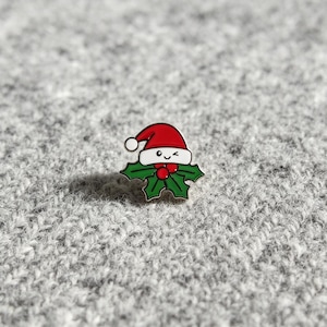 Puede incluir: Un pin de esmalte metálico con forma de rama de acebo con hojas verdes y tres bayas rojas, rematado con un gorro de Papá Noel rojo y blanco. El gorro presenta una cara sonriente con un ojo guiñando, trazada con líneas negras.