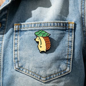 May include: A detailed enamel pin depicting a cartoon hedgehog with a green leaf hat, set against a denim jacket pocket. The hedgehog has a yellow body and brown quills. This charming accessory adds a touch of nature-inspired whimsy to any outfit.