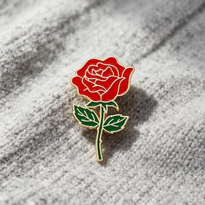 Puede incluir: Un broche de metal dorado y esmalte con forma de rosa individual. La flor está rellena de esmalte rojo vibrante, mientras que el tallo y las dos hojas están rellenos de esmalte verde oscuro. El broche presenta contornos dorados definidos.