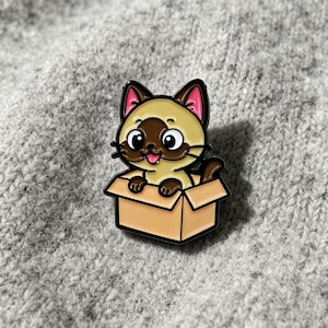 May include: A metal enamel pin featuring a cartoon Siamese cat with cream fur, a dark brown face mask, and pink inner ears. The cat has large black eyes and a small pink tongue sticking out, and it is sitting inside a light brown cardboard box.