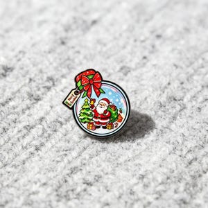 May include: A circular enamel pin featuring a festive snow globe design with a red bow on top. Inside the globe, a cartoon Santa Claus stands next to a decorated Christmas tree and gift boxes. A small rectangular tag attached reads Merry Christmas.