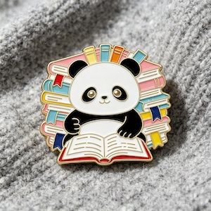 Könnte beinhalten: Eine goldgeränderte Emaille-Anstecknadel mit einem weiß-schwarzen Panda, der vor einem Stapel bunter Bücher sitzt. Der Panda hält ein aufgeschlagenes Buch mit weißen Seiten und roten Rändern. Die Bücher im Hintergrund sind rosa, gelb, blau und orange.